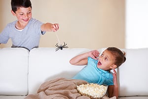 A boy holds a toy spider above another boy eating popcorn on a couch