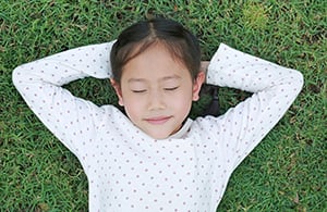 A girl lies on grass with her eyes closed and hands behind her head