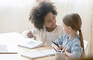 Two girls sit at a table together, one holds a pen and the other a tablet